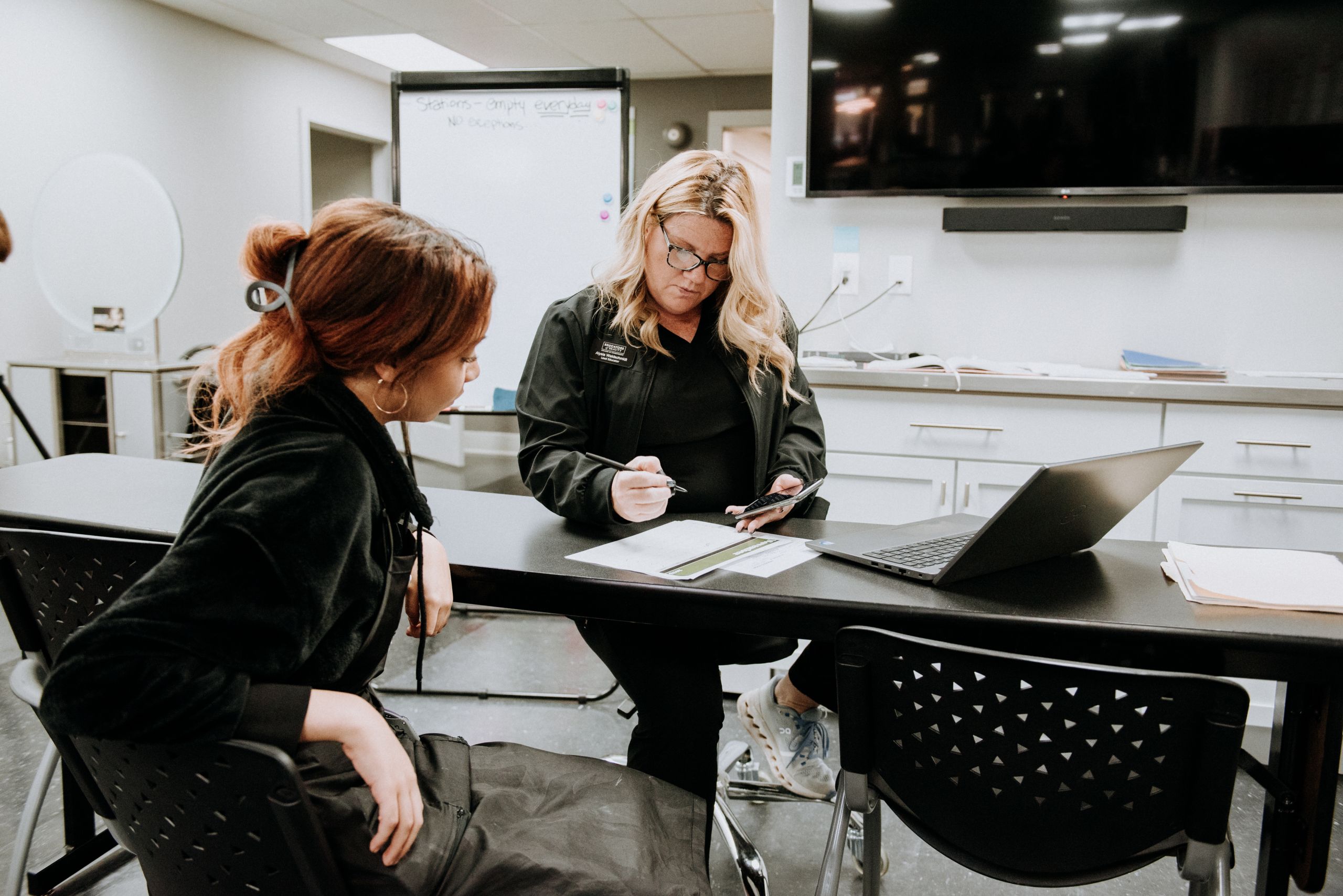 A photo of a woman sitting behind a text explaining something to a woman sitting on the other side of the desk.